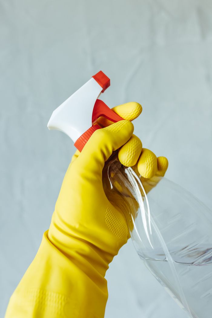 hero-01 A hand wearing a yellow rubber glove holds a red and white spray bottle against a neutral background.